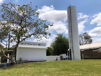 Laboratório do GEHORT - Grupo de Estudos em Horticultura. No topo do prédio os logotipos da UFSCar, Feltrin Sementes, GEHORT, e uma bandeira do Brasil com a frase “Tecnologia UFSCar: Desenvolvendo cultivares tropicalizadas para o Brasil”. Ao lado direito uma caixa d'água alta em formato cilíndrico e a estufa de pimentas ao fundo. Ao lado direito, uma árvore repleta de folhas; no chão, o gramado.